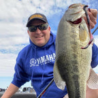 Dustin Wilks wearing a blue Yamaha Pro Fishing hoodie, sunglasses, and a cap holds up a large bass caught on a Culprit Original Worm fishing lure attached to a fish on a boat under a cloudy sky.