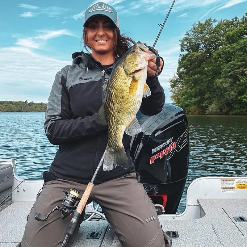 Christina Robinson in a boat on the water holds a large bass fish, wearing a black and gray zip-up jacket, brown pants, and a gray cap. A Mercari Pro XS outboard motor is visible behind her.