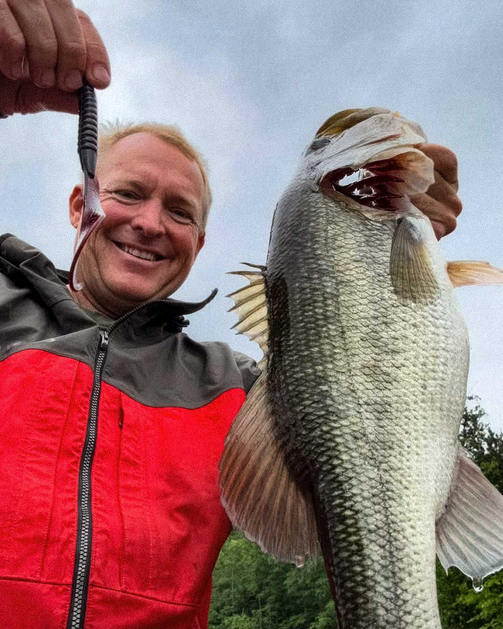 Dustin Wilks in a red and gray zip-up jacket holds up a large bass fish caught with a Culprit Fat Max worm, photographed outdoors against an overcast sky and trees in the background.
