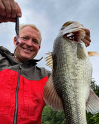 Dustin Wilks in a red and gray zip-up jacket holds up a large bass fish caught with a Culprit Fat Max worm, photographed outdoors against an overcast sky and trees in the background.