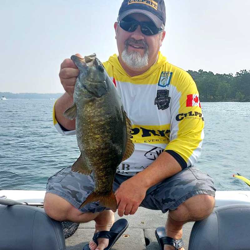 Darren Cavanough wearing a yellow and white Culprit fishing jersey, sunglasses, and a cap sits in a boat on the water, holding up a freshly caught smallmouth bass.