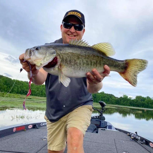Dustin Wilks wearing a black cap with a yellow logo, sunglasses, a dark gray t-shirt, and khaki shorts stands on a boat holding a large bass fish with a Culprit 10-inch Original Worm fishing lure in its mouth. The background shows a calm lake with green trees under an overcast sky.