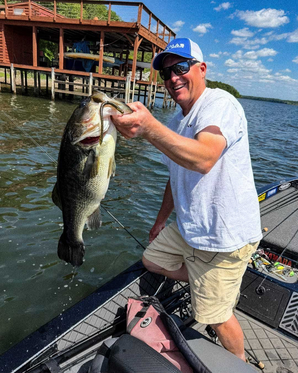 Dustin Wilks wearing a white t-shirt, beige shorts, sunglasses, and a blue cap stands in a boat on a lake, holding up a large bass he just caught with a Culprit T-Rex lure. A wooden dock and water are visible in the background.