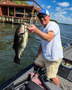 Dustin Wilks wearing a white t-shirt, beige shorts, sunglasses, and a blue cap stands in a boat on a lake, holding up a large bass he just caught with a Culprit T-Rex lure. A wooden dock and water are visible in the background.