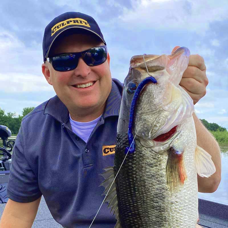 Dustin Wilks wearing a navy blue Culprit cap and matching polo shirt holds a large bass fishing lure on a line outdoors on a boat. The bass was caught on a Culprit Fast Vibe lure.