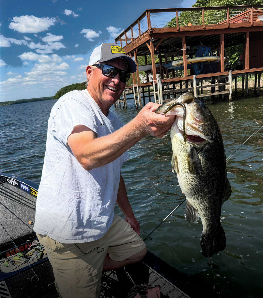 Dustin Wilks in a boat on the water wearing a white t-shirt and khaki shorts holds up a large bass fish he caught with a Culprit T-Rex finesse worm. A wooden dock is visible in the background under a blue sky with clouds.