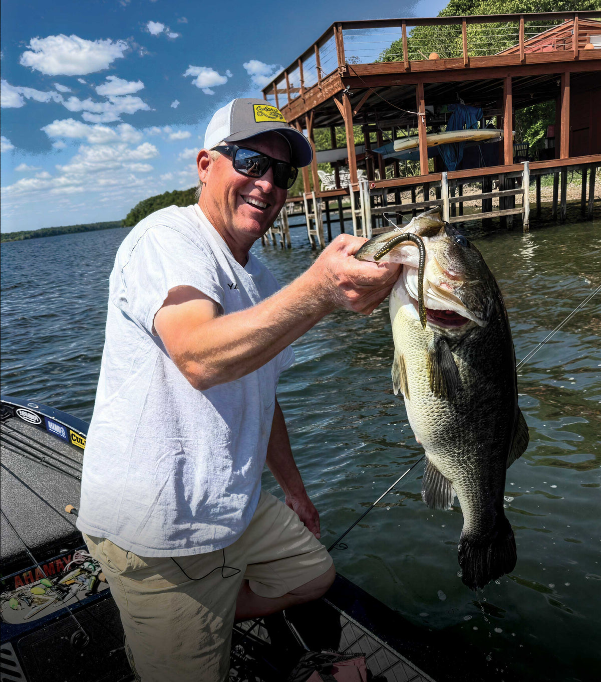 Dustin Wilks in a boat on the water wearing a white t-shirt and khaki shorts holds up a large bass fish he caught with a Culprit T-Rex finesse worm. A wooden dock is visible in the background under a blue sky with clouds.