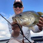 Dustin Wilks wearing a brown Falcon cap and sunglasses holds a large Culprit Fat Max lure attached to a hook while holding a freshly caught largemouth bass in a boat under a blue sky.