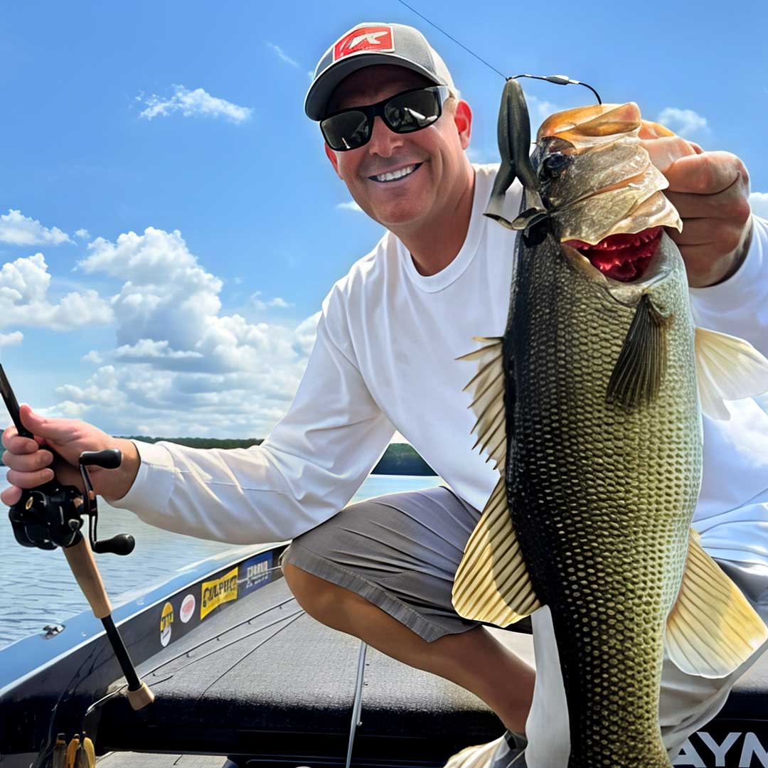 A smiling Dustin Wilks in a white long-sleeve shirt, gray cap, and sunglasses sits in a boat on the water, holding up a large bass fish hooked on the Culprit Incredi-Frog fishing lure.