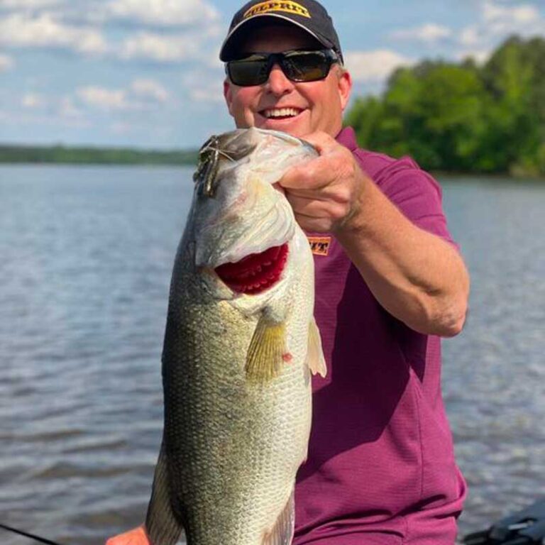 Dustin Wilks wearing sunglasses and a black Culprit cap holds up a large bass fish on a boat on the water, with trees and blue sky in the background.