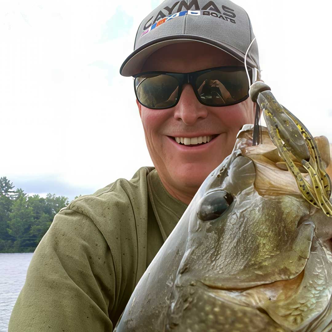 Dustin Wilks wearing a gray Caymas Waterfowl cap and sunglasses holds a large fish with a Culprit Shakin Neko Craw soft plastic lure in his mouth, outdoors near water and trees.