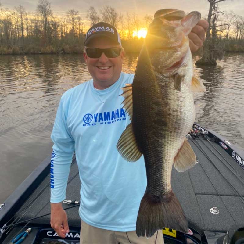 Dustin Wilks on a boat wearing a light blue Yamaha Pro Fishing long-sleeve shirt and a black cap holds up a large bass fish during sunset over a lake.
