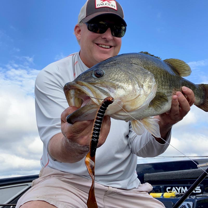 Dustin Wilks wearing a brown and white Halyard cap, sunglasses, and a white long-sleeve shirt holds a largemouth bass in a boat while dangling a Crawdad Fat Max soft plastic worm lure.