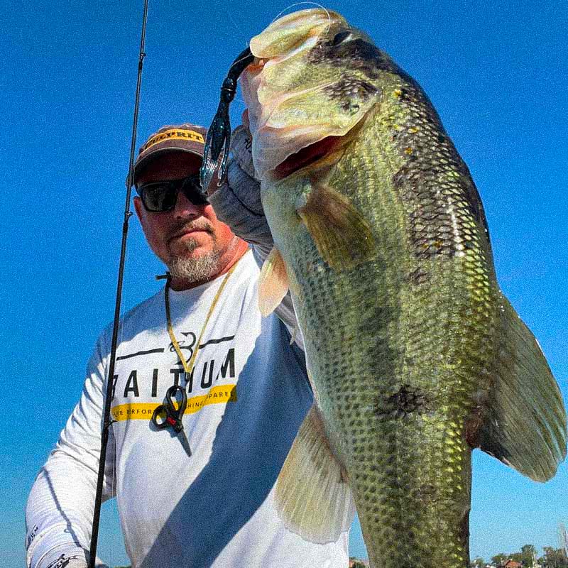 Matt Koser (aka Get Ugly Fishing) wearing a white long-sleeve shirt with a yellow and black 'PLATINUM' logo and a Culprit ball cap holds up a large bass caught on a Culprit Shakin Neko Craw on a fishing rod against a bright blue sky.
