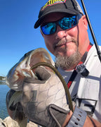 Matt Koser wearing a black cap with a yellow and red logo, blue mirrored sunglasses, and a white t-shirt holds a largemouth bass in a gray silicone glove while demonstrating a Culprit T-Rex soft plastic fishing lure. The background shows a bright blue sky and water.