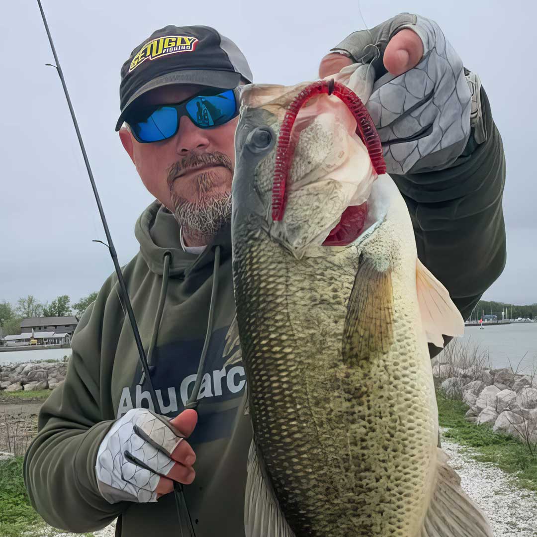 Matt Koser wearing a green hoodie, a black and yellow GetUglyFishing cap, and blue sunglasses holds a large bass and T-Rex fishing lure on a fishing rod outdoors by the water.