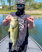 Nic Howell on a boat wearing a black cap and face mask holds up a largemouth bass while showing a Red Shad Culprit Original Worm fishing lure on a hook.