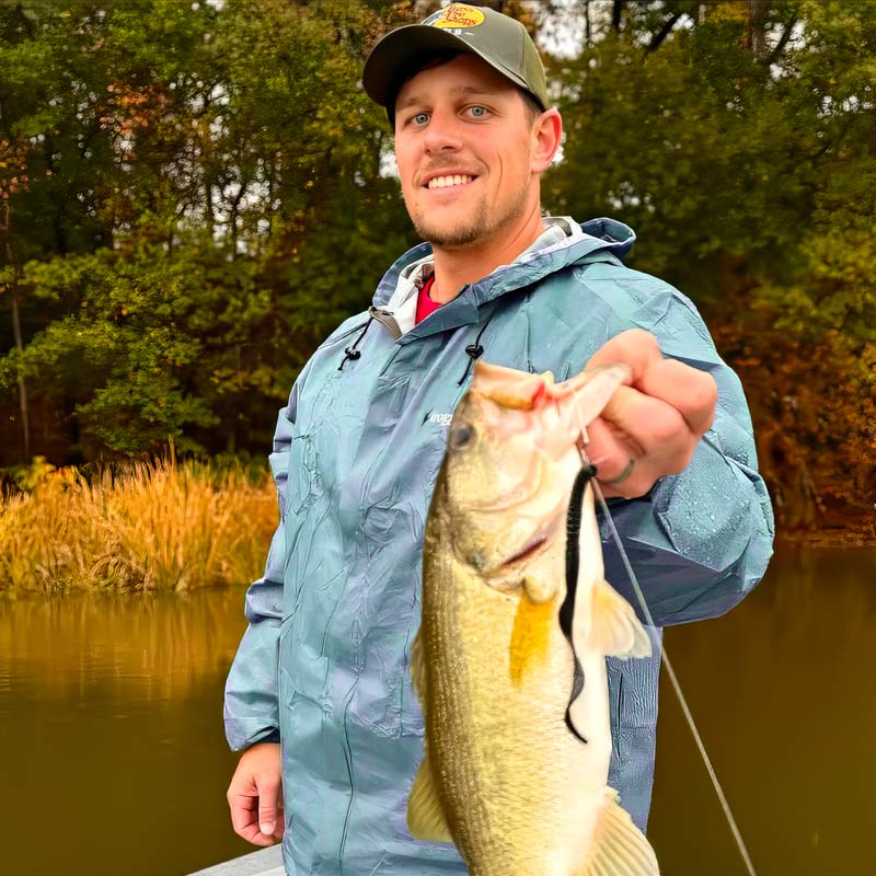 Justin Alford wearing a blue rain jacket and a green cap holds a largemouth bass on a fishing line outdoors by a lake with trees in the background.