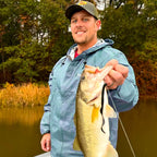 Justin Alford wearing a blue rain jacket and a green cap holds a largemouth bass on a fishing line outdoors by a lake with trees in the background.