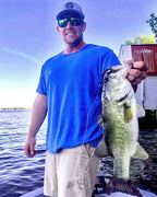 Justin Alford wearing a blue t-shirt, beige shorts, sunglasses, and a baseball cap stands on a boat holding a large freshly caught largemouth bass over the side of the boat, with water and a house in the background.