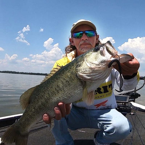 Jonathan Mickler kneeling in a boat on the water, holding a large bass fish with a Culprit Original Worm soft plastic lure in his mouth. He is wearing a yellow and white shirt, blue jeans, and sunglasses under a blue sky with white clouds.