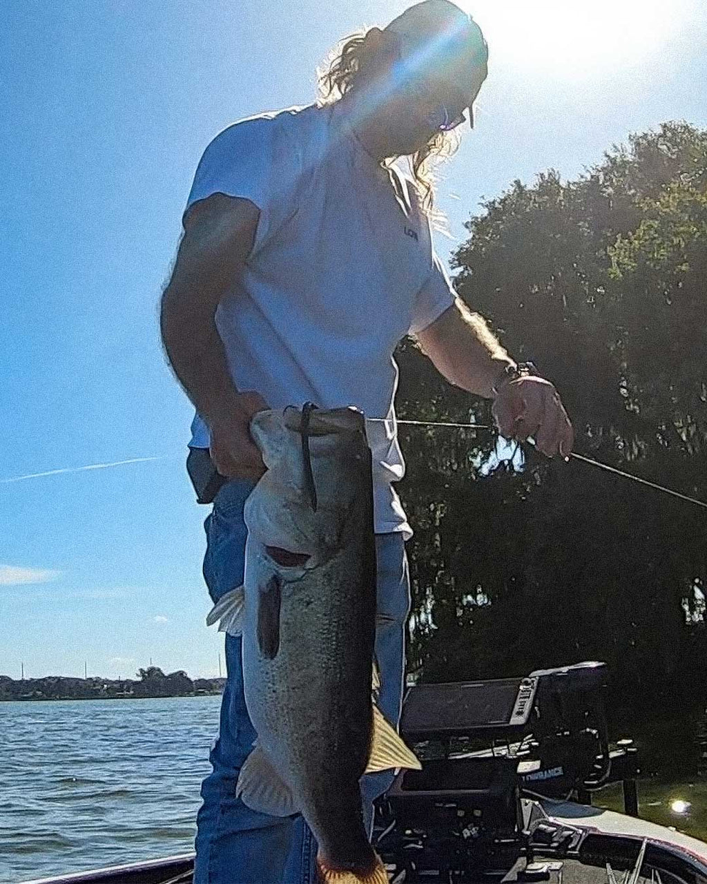 Jonathan Mickler on a boat holds up a large fish he just caught on a Culprit T-Rex lure while wearing a white t-shirt, blue jeans, and sunglasses, with trees and water in the background under bright sunlight.