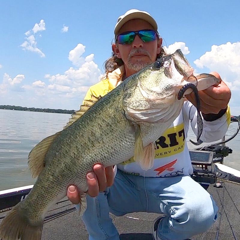 Jonathan Mickler kneeling on a boat holds a large bass fish with a Culprit Original Worm fishing lure in its mouth. The man is wearing a beige cap, sunglasses, a yellow shirt, and a white long-sleeve shirt with a Culprit logo.