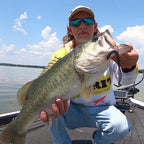 Jonathan Mickler kneeling on a boat holds a large bass fish with a Culprit Original Worm fishing lure in its mouth. The man is wearing a beige cap, sunglasses, a yellow shirt, and a white long-sleeve shirt with a Culprit logo.
