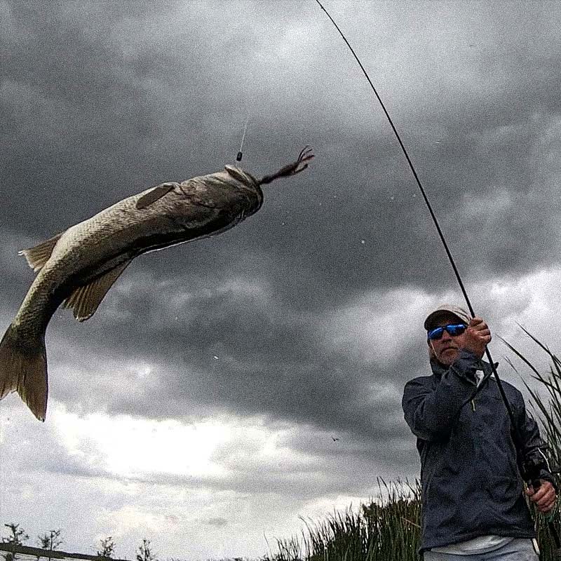 Jonathan Mickler wearing sunglasses and a dark jacket near a grassy shore holding a fishing rod, with a large fish caught with a Culprit Shakin Neko Craw leaping through the air toward the boat under a dramatic cloudy sky.