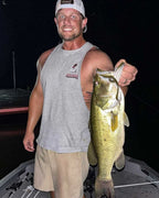 Justin Warren in a gray tank top and khaki shorts holds a llargemouth bass freshly caught on a Culprit Original Worm lure at night while standing in a boat.