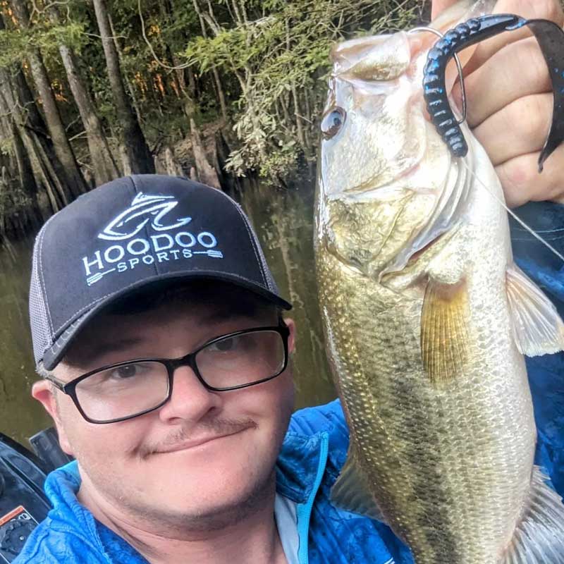 Zach Peeples wearing a black HOODOO SPORTS cap and glasses holds up a freshly caught largemouth bass with a Culprit Fat Max lure in its mouth, set against a background of a swamp with cypress trees.