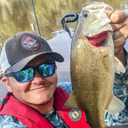 Zach Peeples wearing a gray and black baseball cap with a circular logo, sunglasses, and a red life jacket holds up a freshly caught largemouth bass with a black Culpit Fat Max fishing lure in its mouth, with a lake in the background.
