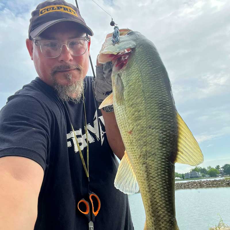 Matt Koser wearing a black t-shirt and a cap with 'CULPRIT' on it holds up a large bass fish caught on a Culprit Shakin Neko Craw outdoors near the water under a cloudy sky.