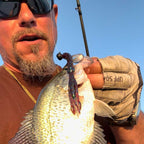 Matt Koser wearing sunglasses and a tan fishing glove holds a freshly caught fish with a soft plastic Culprit Shakin Neko Craw attached, set against a bright blue sky.