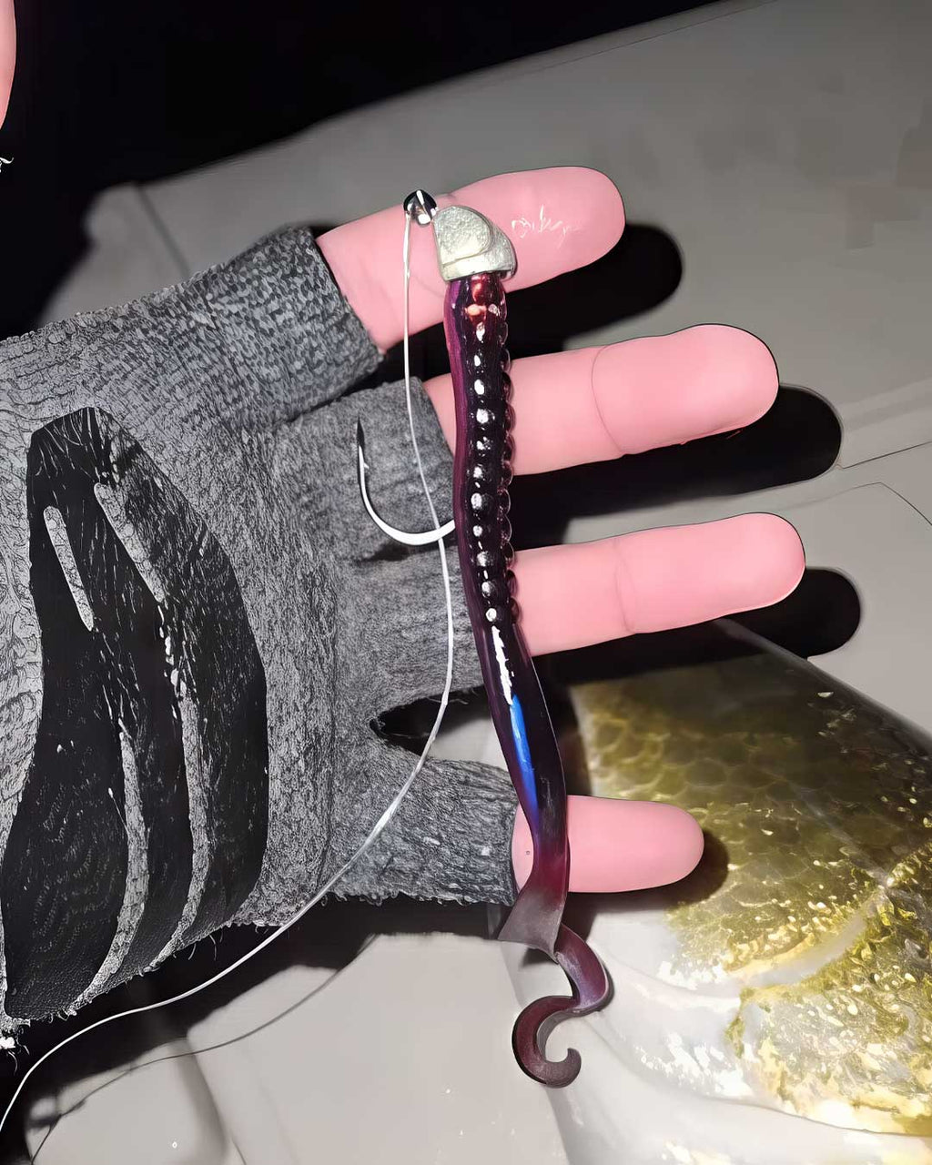 Sawyer Martin holds a fishing lure being held on a line over a fish, with a gloved hand visible. The Culprit Original Worm lure has a silver top, a dark purple segmented body with iridescent blue-purple highlights, and a hook at the bottom.