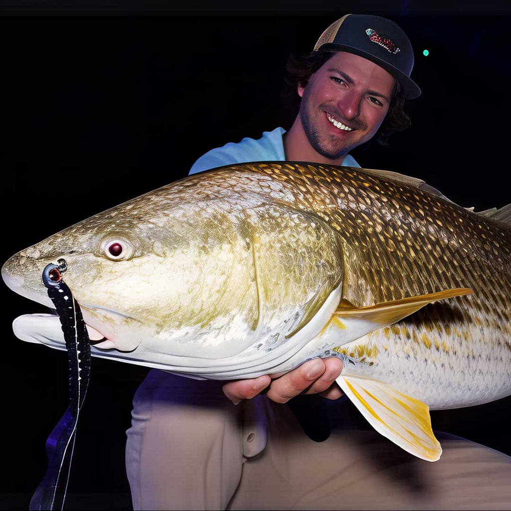 A smiling Sawyer Martin in a blue shirt and tan pants holds a very large redfish with a Culprit Original worm fishing lure in its mouth against a dark background.