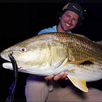 A smiling Sawyer Martin in a blue shirt and tan pants holds a very large redfish with a Culprit Original worm fishing lure in its mouth against a dark background.
