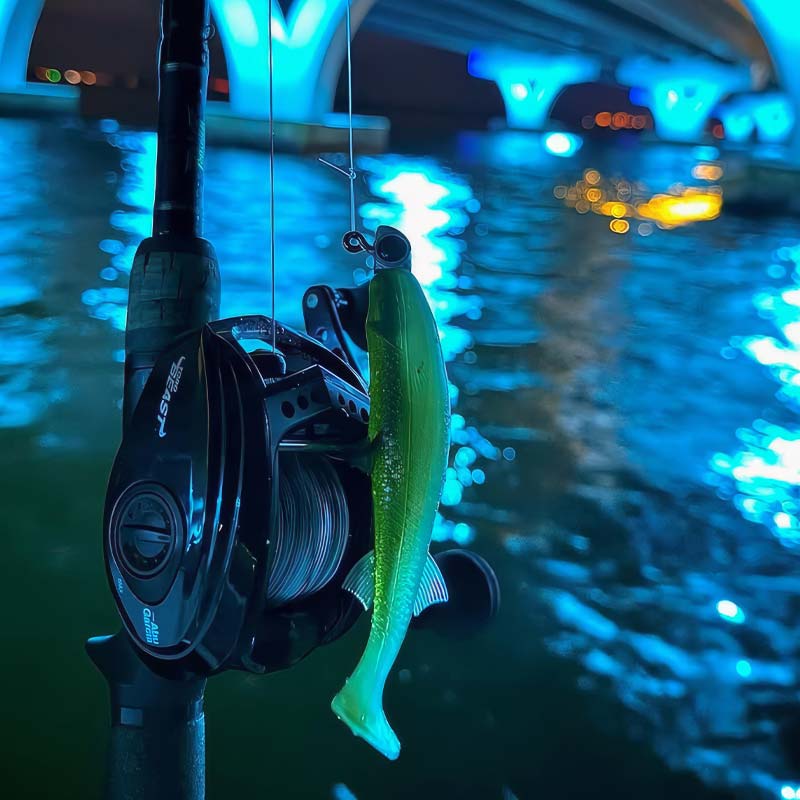 A fishing rod with a black spinning reel mounted on a pole, holding a bright green Riptide Mullet soft plastic fishing lure, photographed outdoors at night over water with a lit bridge in the background.