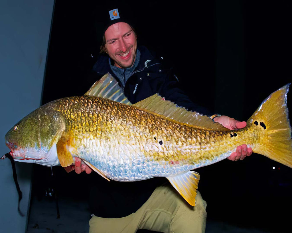 Sawyer Martin in a black beanie and dark jacket holds a very large redfish against a dark nighttime background.