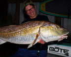 Sawyer Martin wearing glasses and a cap holds a very large redfish caught on a Riptide Mullet over his shoulder in a nighttime setting. A white boat trailer with the brand BÖTE is visible in the background, along with a paddle board. 