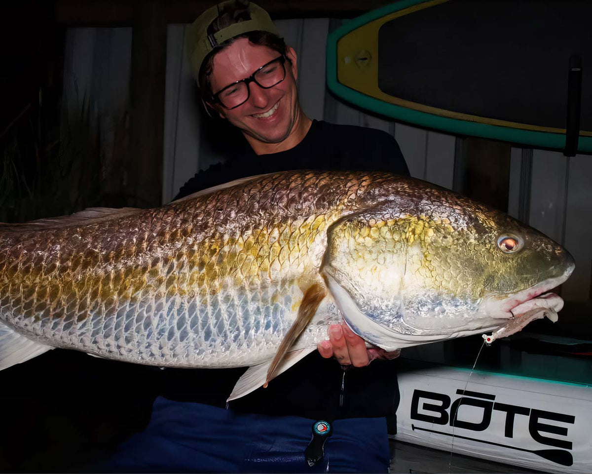 Sawyer Martin wearing glasses and a cap holds a very large redfish caught on a Riptide Mullet over his shoulder in a nighttime setting. A white boat trailer with the brand BÖTE is visible in the background, along with a paddle board. 