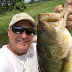 Dustin Wilks, wearing a beige cap, sunglasses, and a white t-shirt holds up a large freshly caught bass fish in an outdoor setting by a river or lake, with green grassy hills in the background.