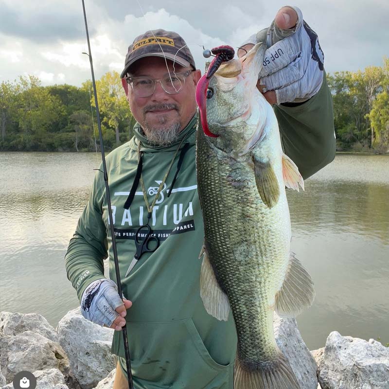 Matt Koser wearing a green Nautica hoodie and a camouflage cap holds up a large bass fish with a red lip fishing lure, standing on rocks by a lake.