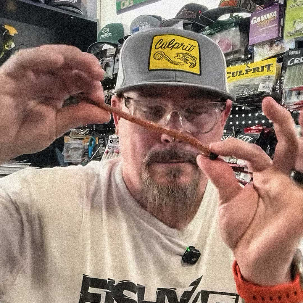 Matt Koser wearing a gray Culpit brand cap and a cream-colored t-shirt holds a T-Rex fishing lure in front of his face inside what appears to be a retail store.